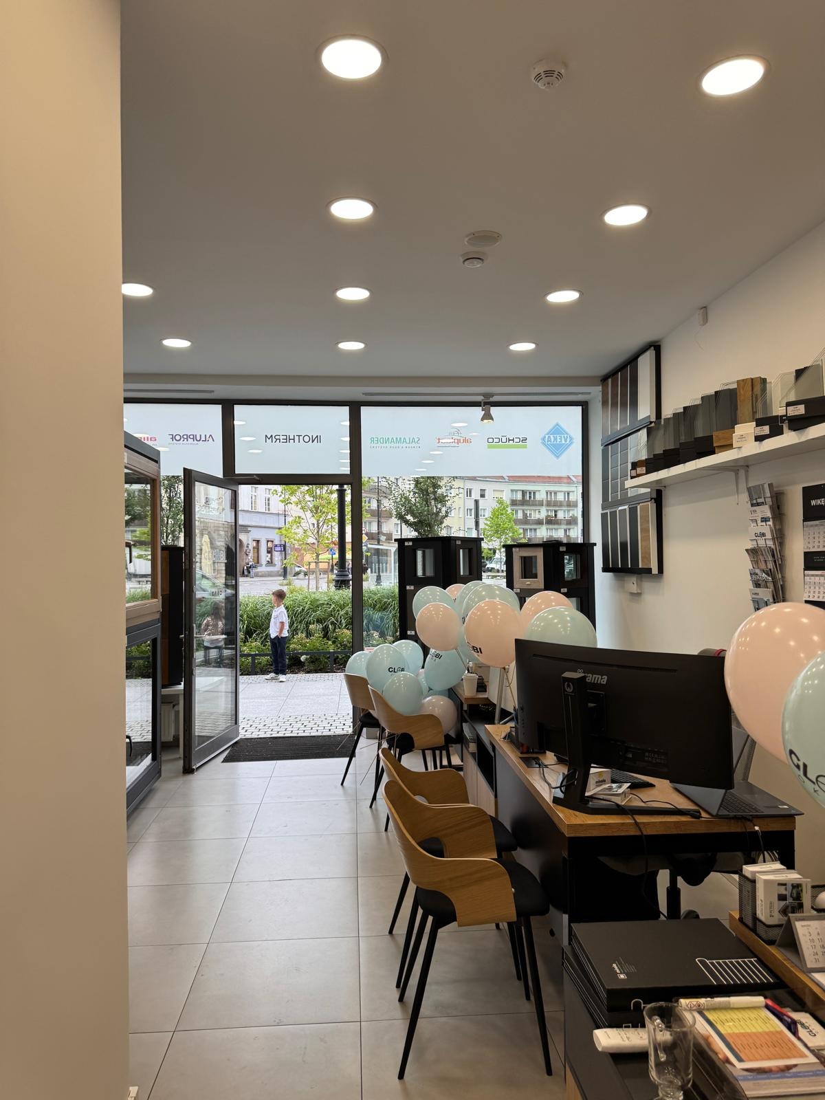 Salon z ekspozycją okien i drzwi, dekorowany balonami. Widoczne próbki profili okiennych i drzwiowych na ścianie. Na zewnątrz, przez otwarte drzwi, widać fragment ulicy.