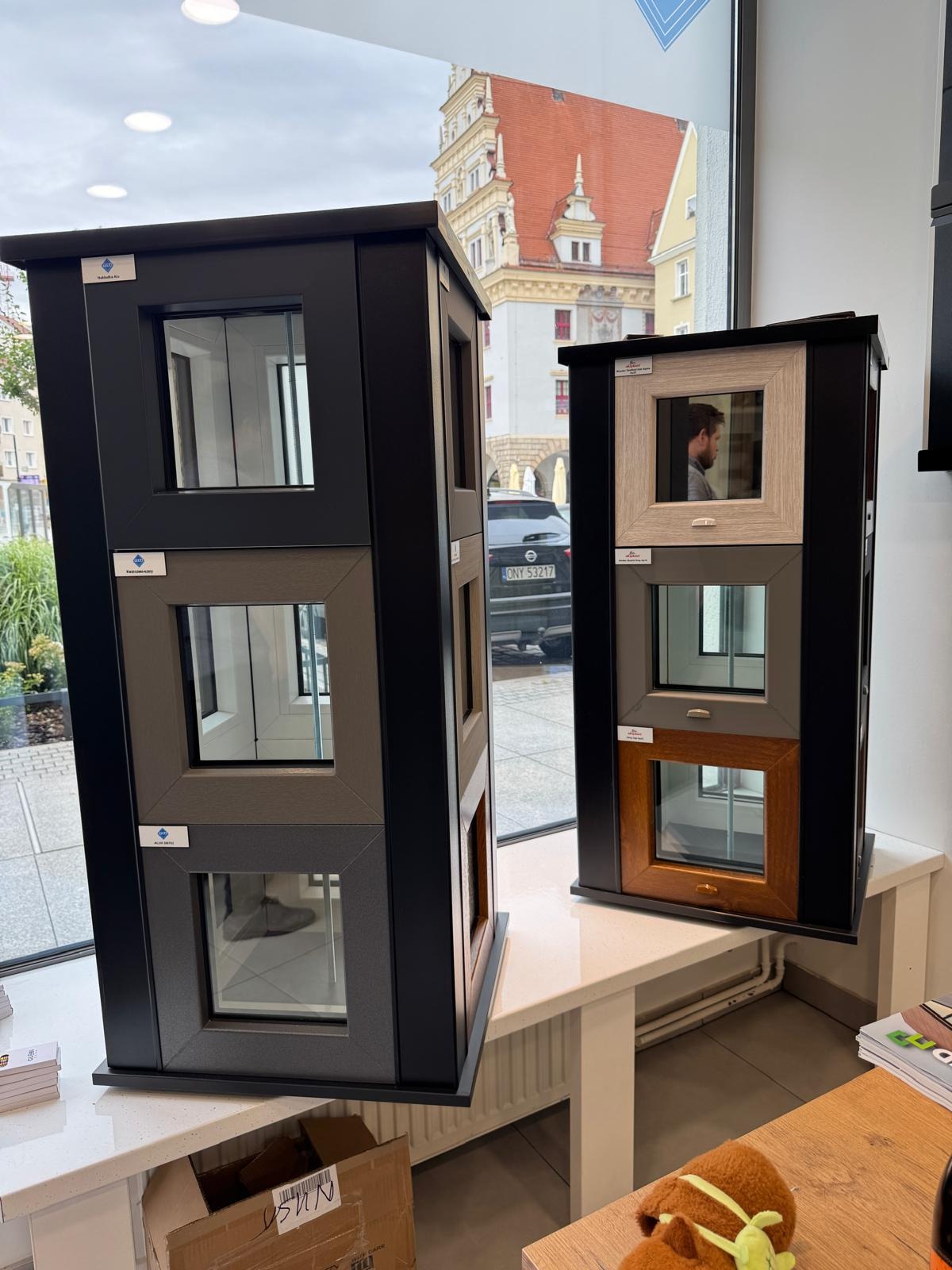 Ekspozycja okien z różnymi ramami i wykończeniami w salonie, na tle okna z widokiem na zabytkową architekturę miejską. Prezentacja wzornictwa i kolorystyki.