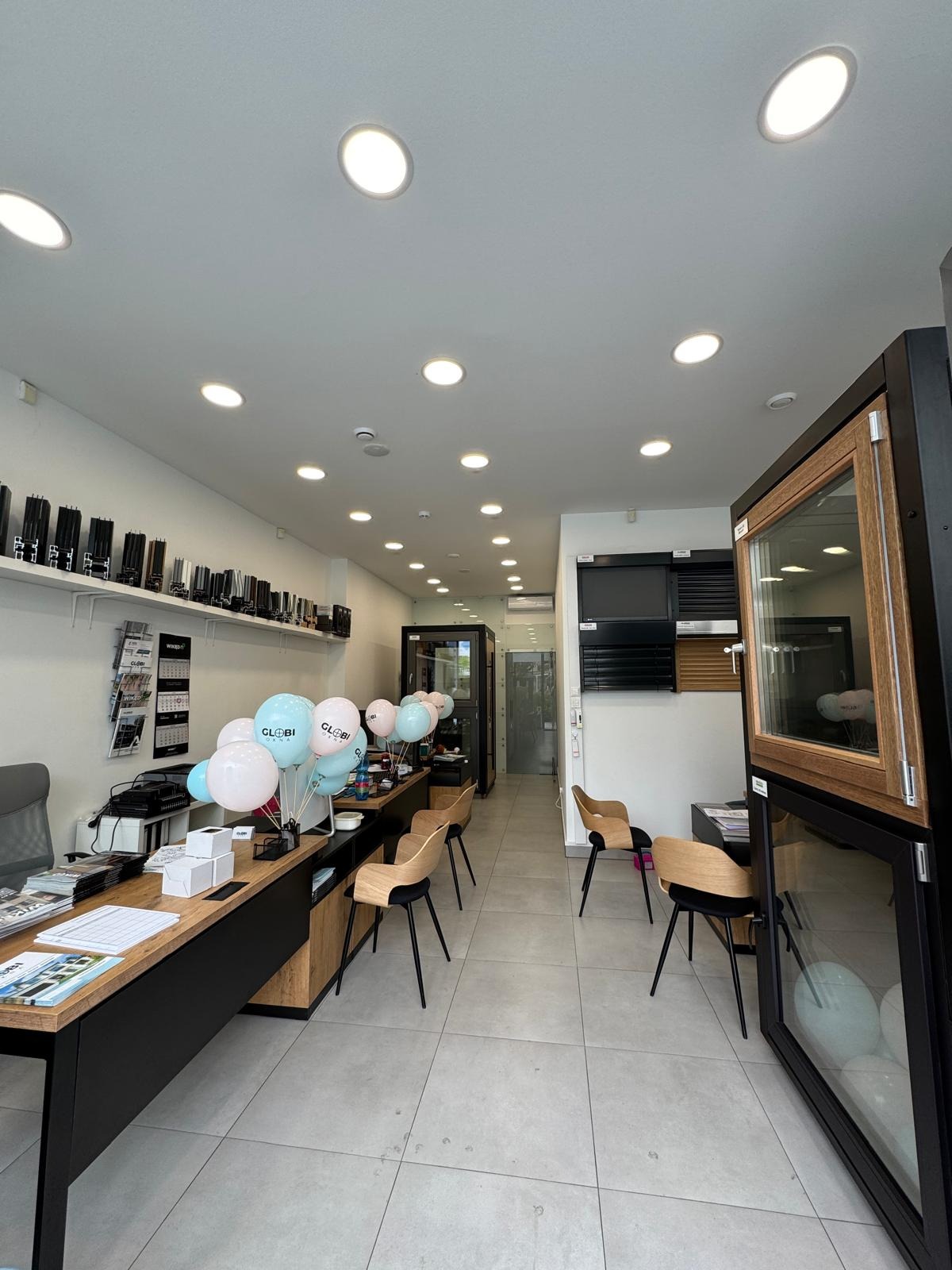 Wnętrze salonu z ekspozycją okien. Widoczne biurko z dekoracjami, krzesła oraz próbki okien z różnymi wykończeniami. Jasne oświetlenie i minimalistyczny design.