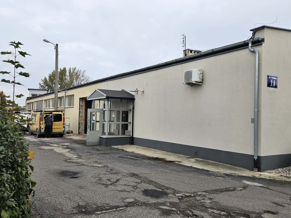 Budynek z przeszklonym wejściem i zaparkowanym żółtym busem. Widoczny klimatyzator na ścianie i tabliczka z adresem. Pochmurne niebo w tle.