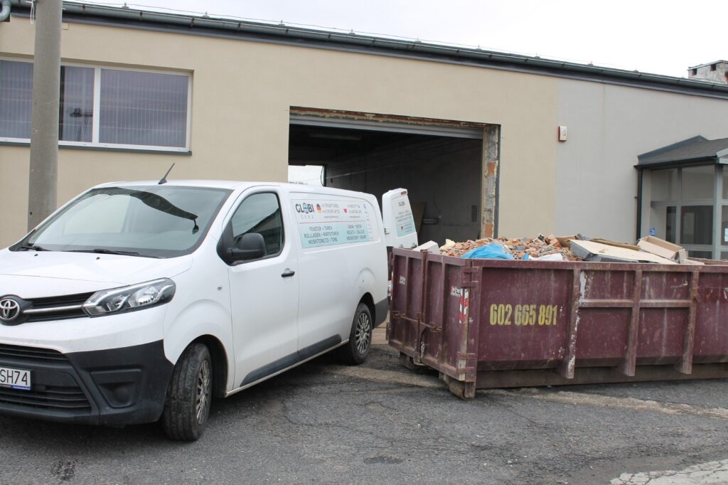 Kontener z gruzem po demontażu, biały van firmy z reklamą okien i rolet na tle budynku z otwartym wjazdem. Widoczne okno z kratami.