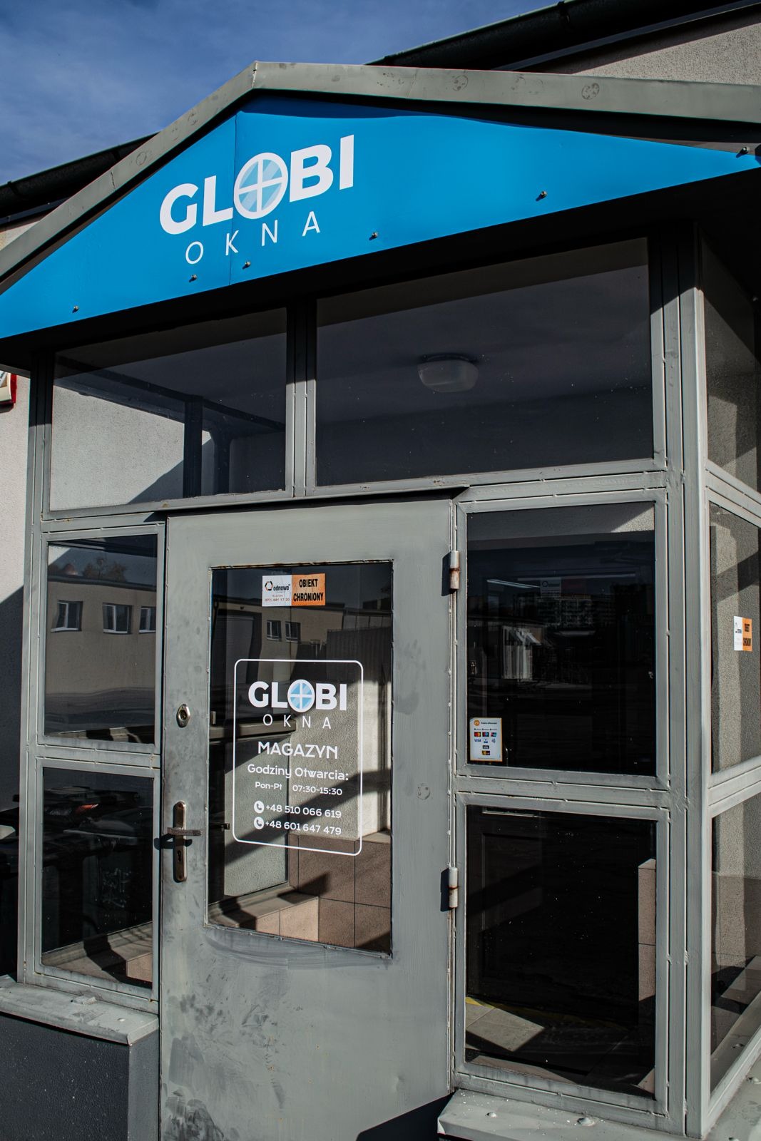 Wejście do salonu z oknami marki Globi Okna, szara rama, przeszklone drzwi z naklejką informacyjną, niebieski daszek z logo, widok z zewnątrz, dzień.