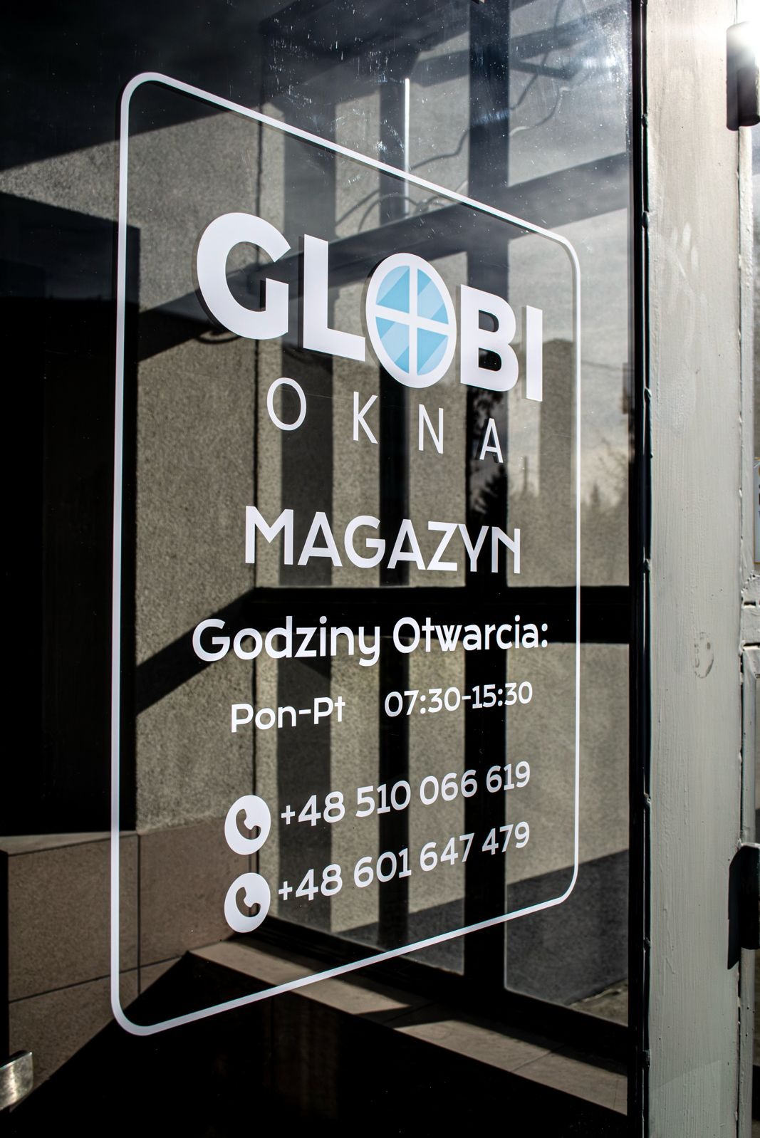Szyba z napisem 'GLOBI OKNA MAGAZYN' i godzinami otwarcia, widoczne numery telefonów. Cień na ścianie. Ujęcie z bliska, ostre światło.