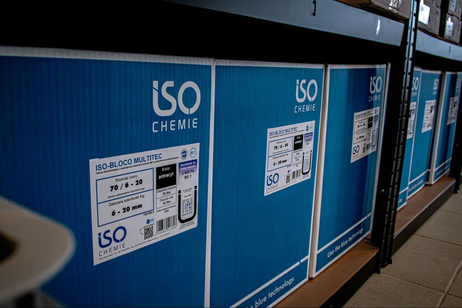 Rząd niebieskich kartonów ISO Chemie z etykietami informacyjnymi na metalowym regale. Widoczne rozmiary, kolory i certyfikaty. Magazyn materiałów budowlanych.