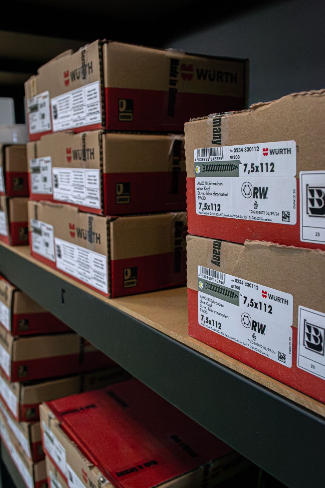 Półka magazynowa z kartonami śrub Wurth 7,5x112. Etykiety z detalami produktu widoczne na pierwszym planie. Magazynowe oświetlenie.