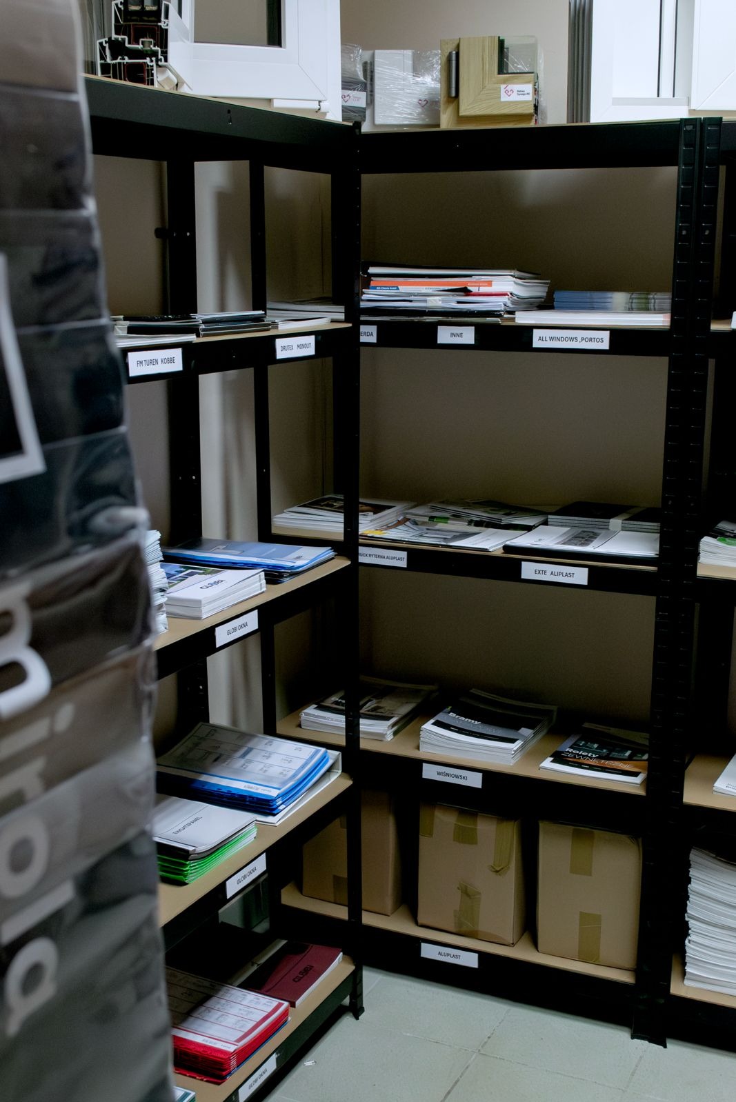 Regał magazynowy z próbkami i katalogami okien, drzwi i bram garażowych różnych producentów, oświetlony neutralnym światłem.