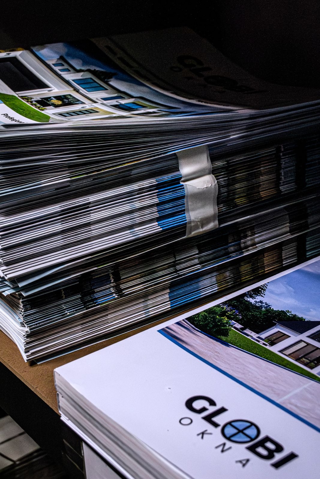 Stos katalogów z oknami i drzwiami firmy Globi Okna, zabezpieczony taśmą, leżący na blacie. Widoczne zdjęcia domów z zamontowanymi oknami.