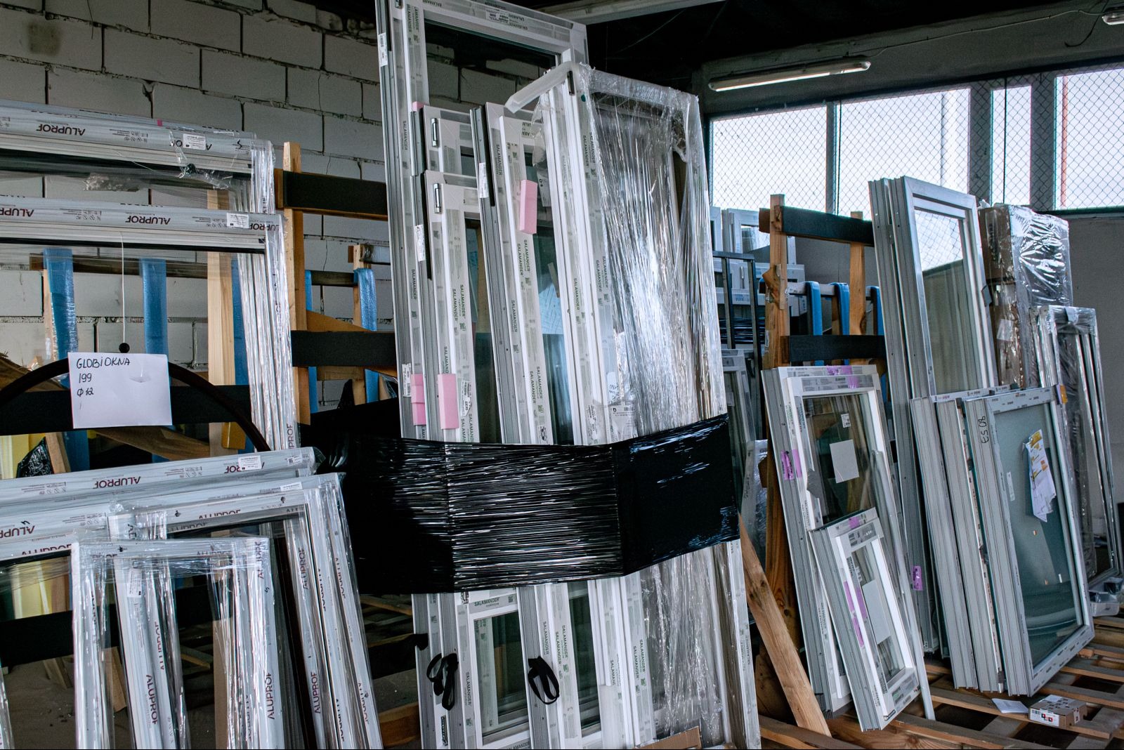 Magazyn okien i ram okiennych różnych producentów (Aluprof, Salamander) składowanych na drewnianych paletach. Część zafoliowana, widoczne etykiety i notatki.