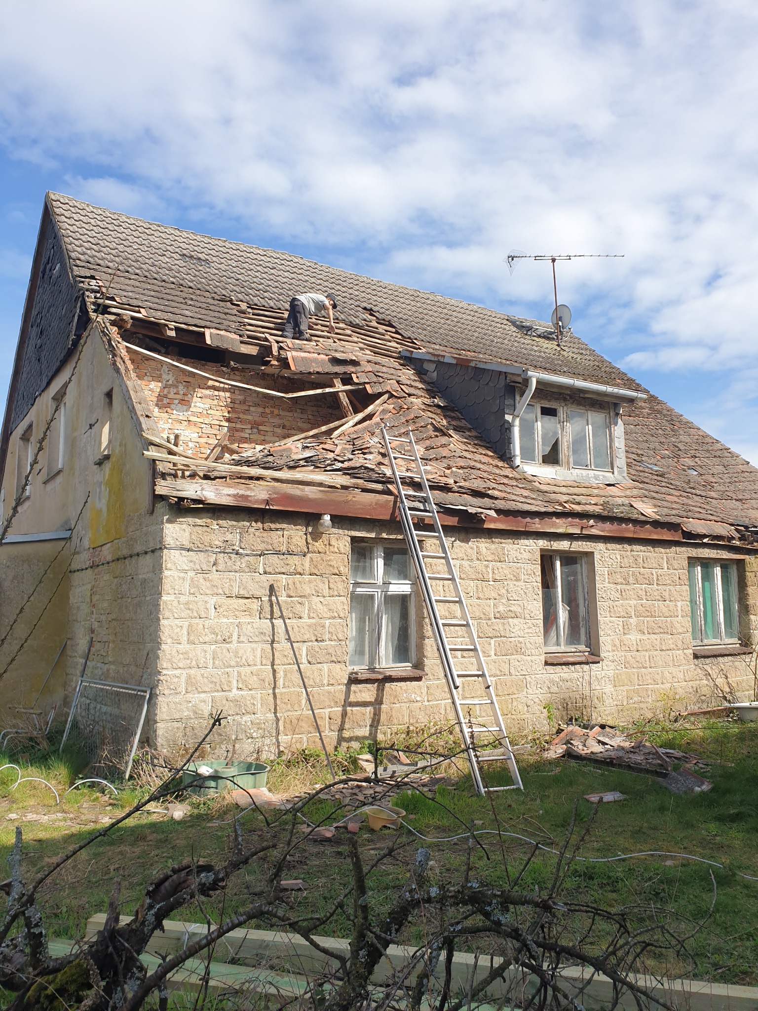 Dom z częściowo usuniętym, starym dachem z dachówki, drabina oparta o ścianę, pracownik na dachu. Widoczne uszkodzenia więźby i ceglana ściana szczytowa.