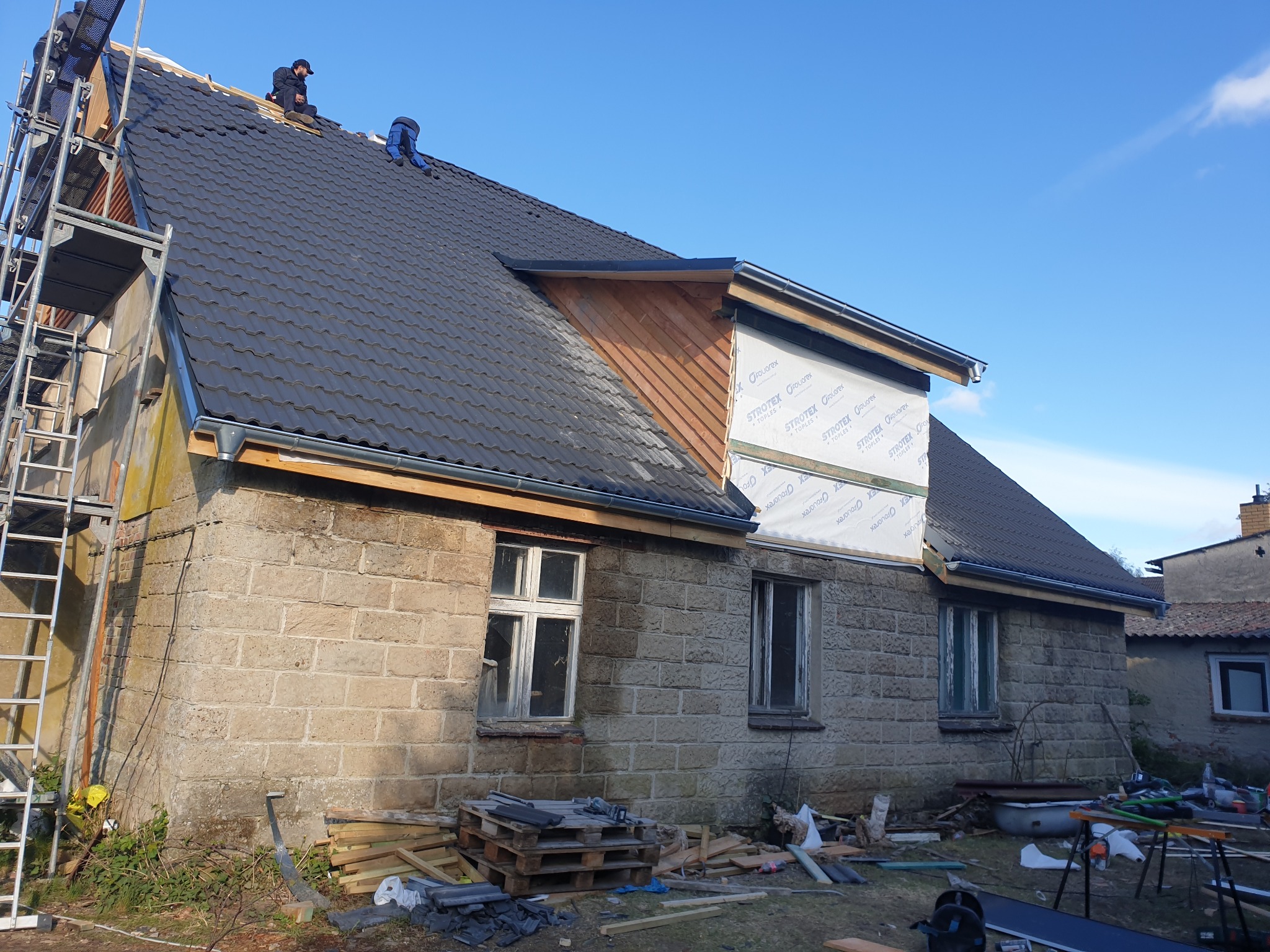 Dom z częściowo wymienionym pokryciem dachu, widoczne rusztowanie i pracownicy na dachu. Część dachu z membraną, przygotowana do dalszych prac.