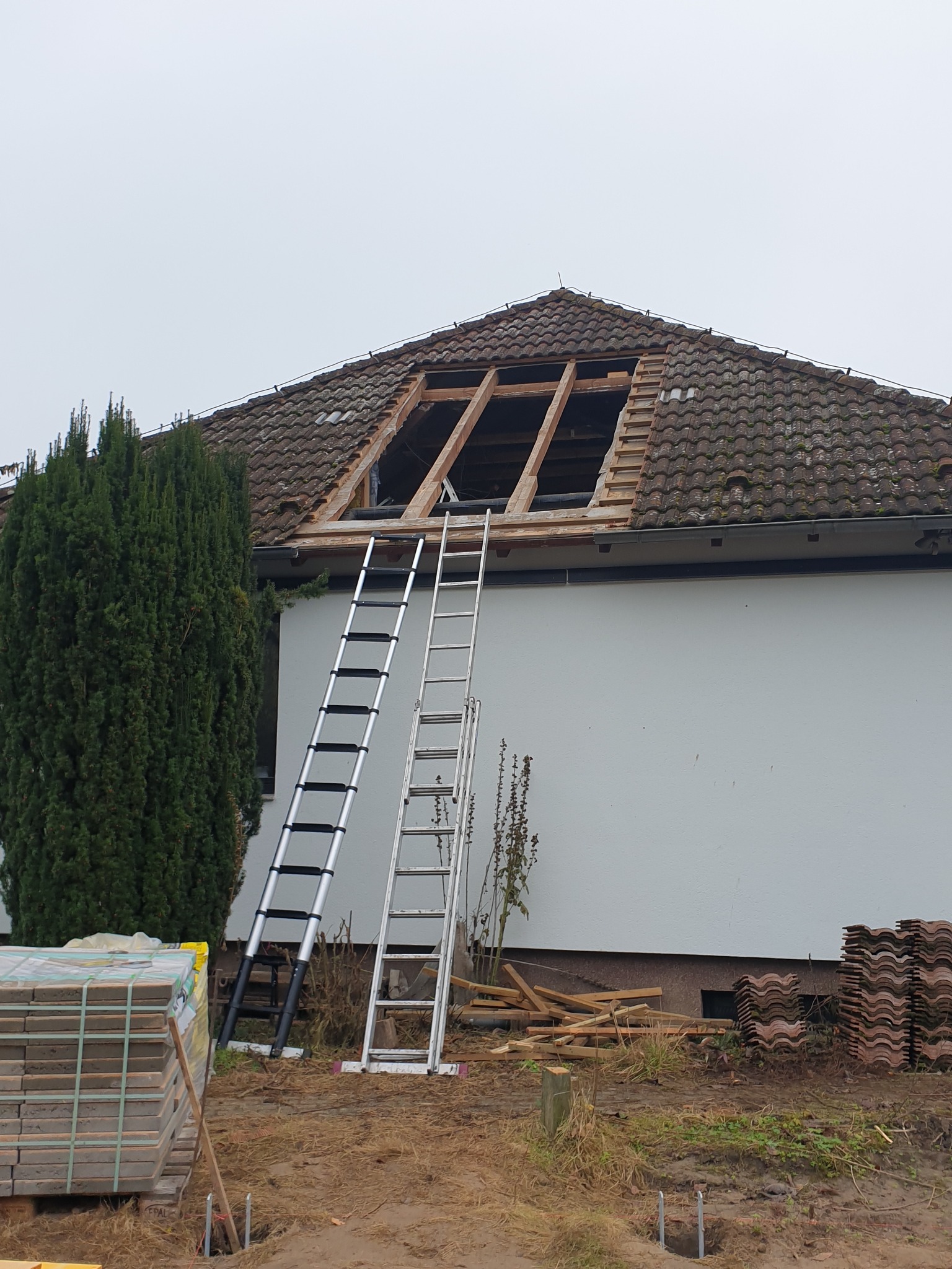 Dom z częściowo zdjętym dachem, widoczne krokwie. Dwie drabiny opierają się o ścianę. Obok leżą dachówki w paletach i stosach. Etap prac dekarskich.