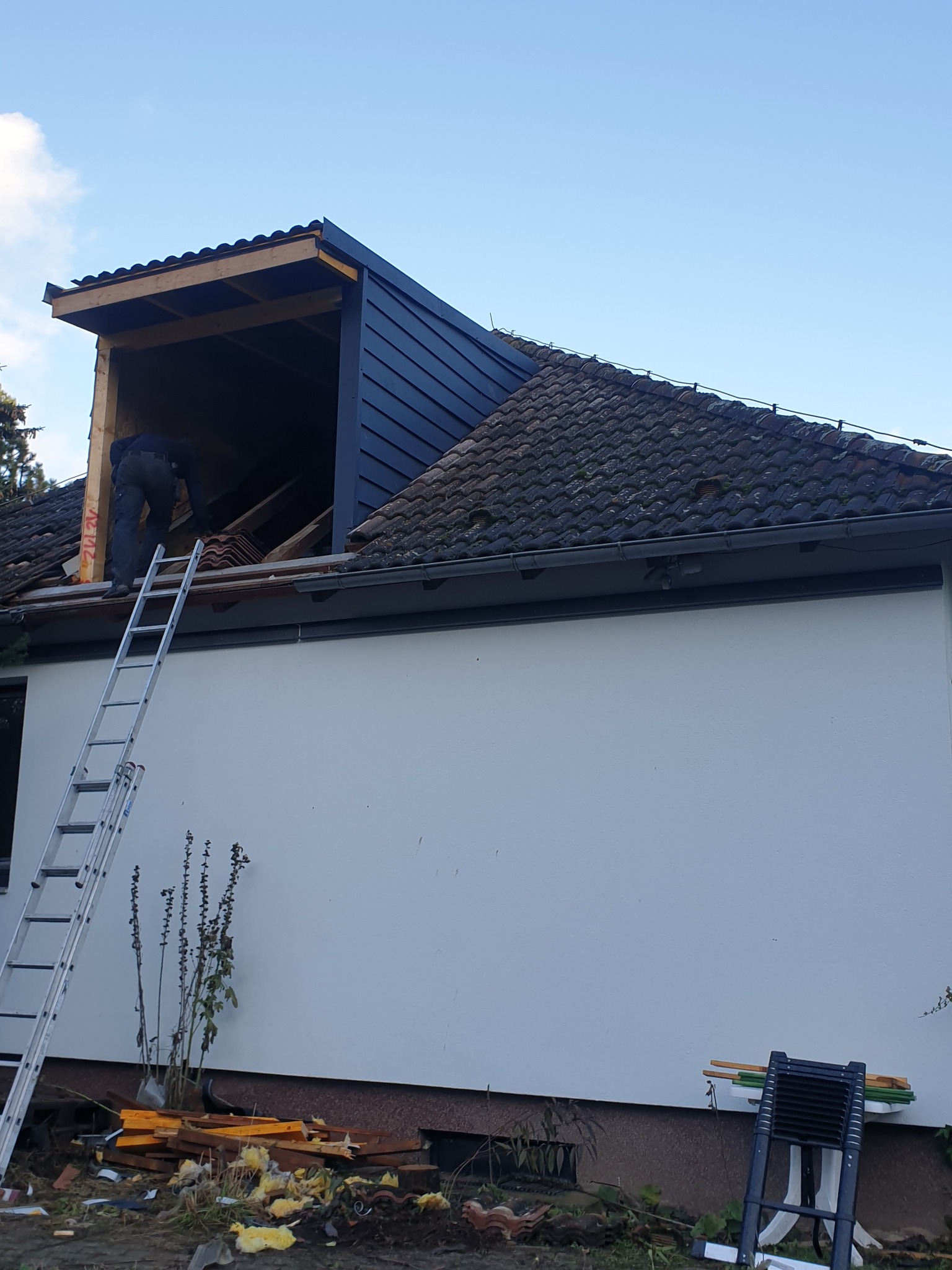 Prace dekarskie: mężczyzna na drabinie układa dachówkę na dobudowanej części dachu. Widoczne elementy konstrukcyjne i fragment starego dachu z rynną.