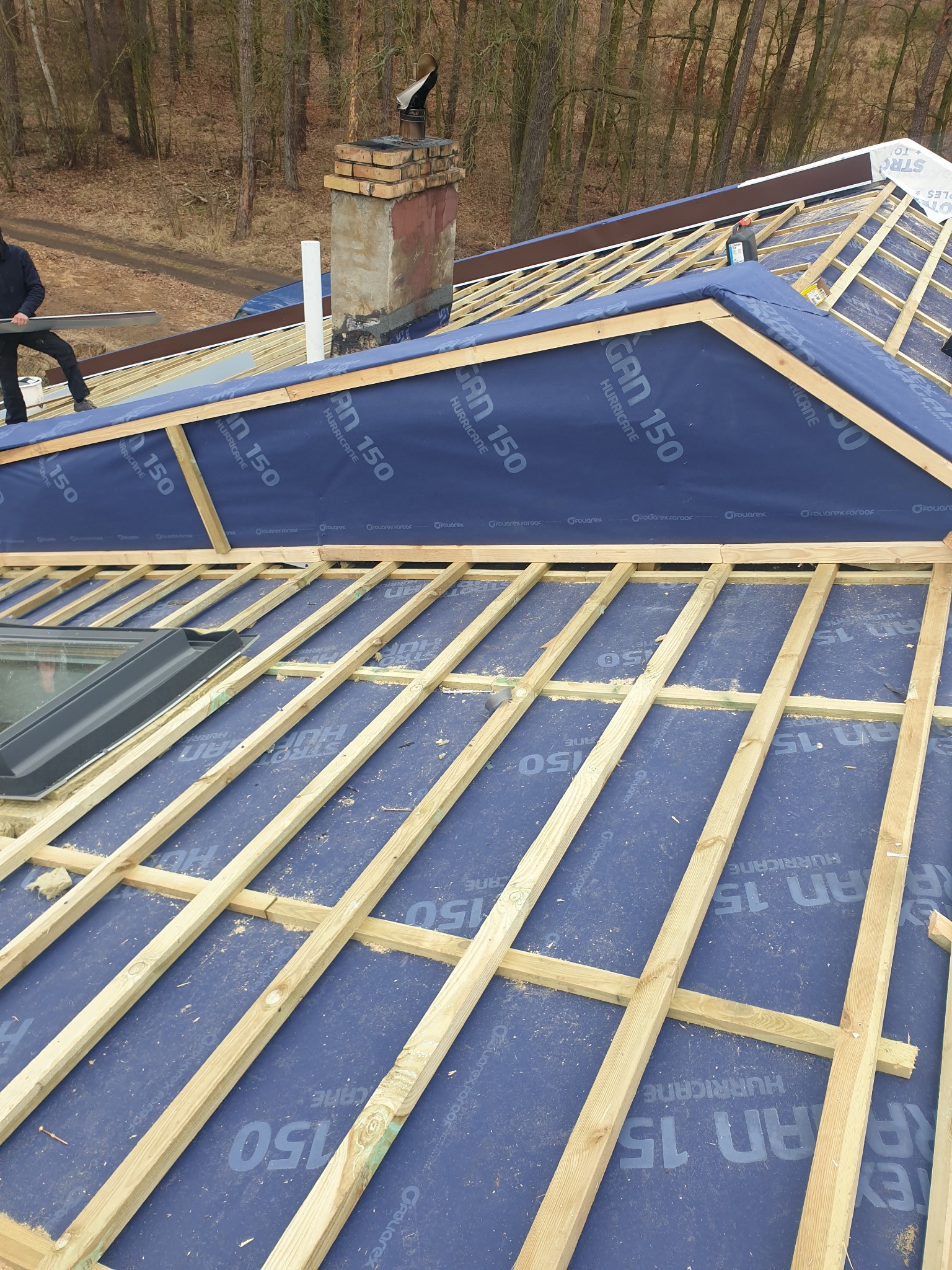 Konstrukcja dachu w trakcie budowy: drewniane łaty na membranie, okno dachowe i komin w tle. Widoczny fragment sylwetki dekarza z narzędziem.