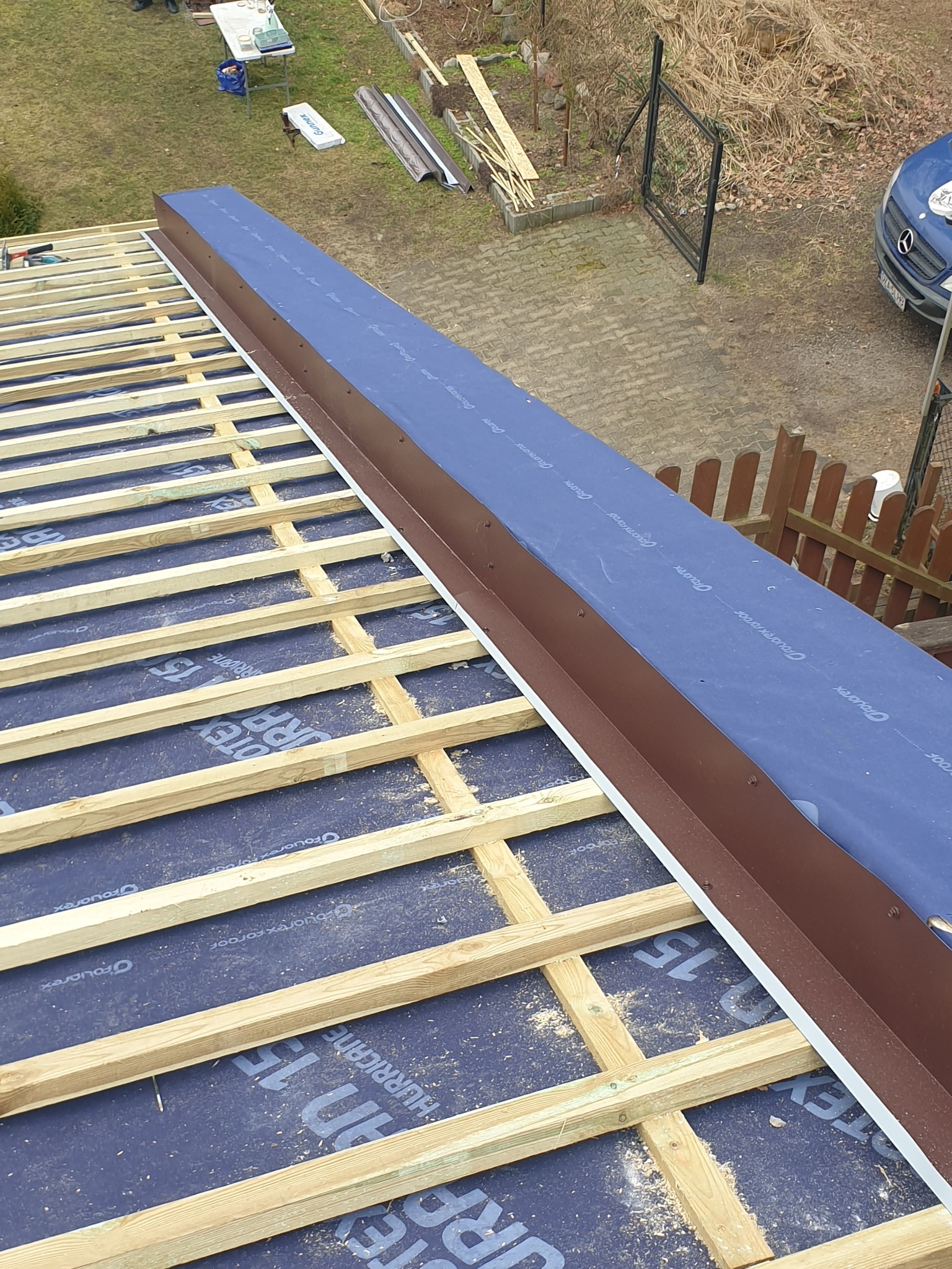 Konstrukcja dachu z membraną i obróbką blacharską w kolorze brązowym, widok z góry. Elementy drewniane, niebieska membrana i blacha tworzą regularny wzór.