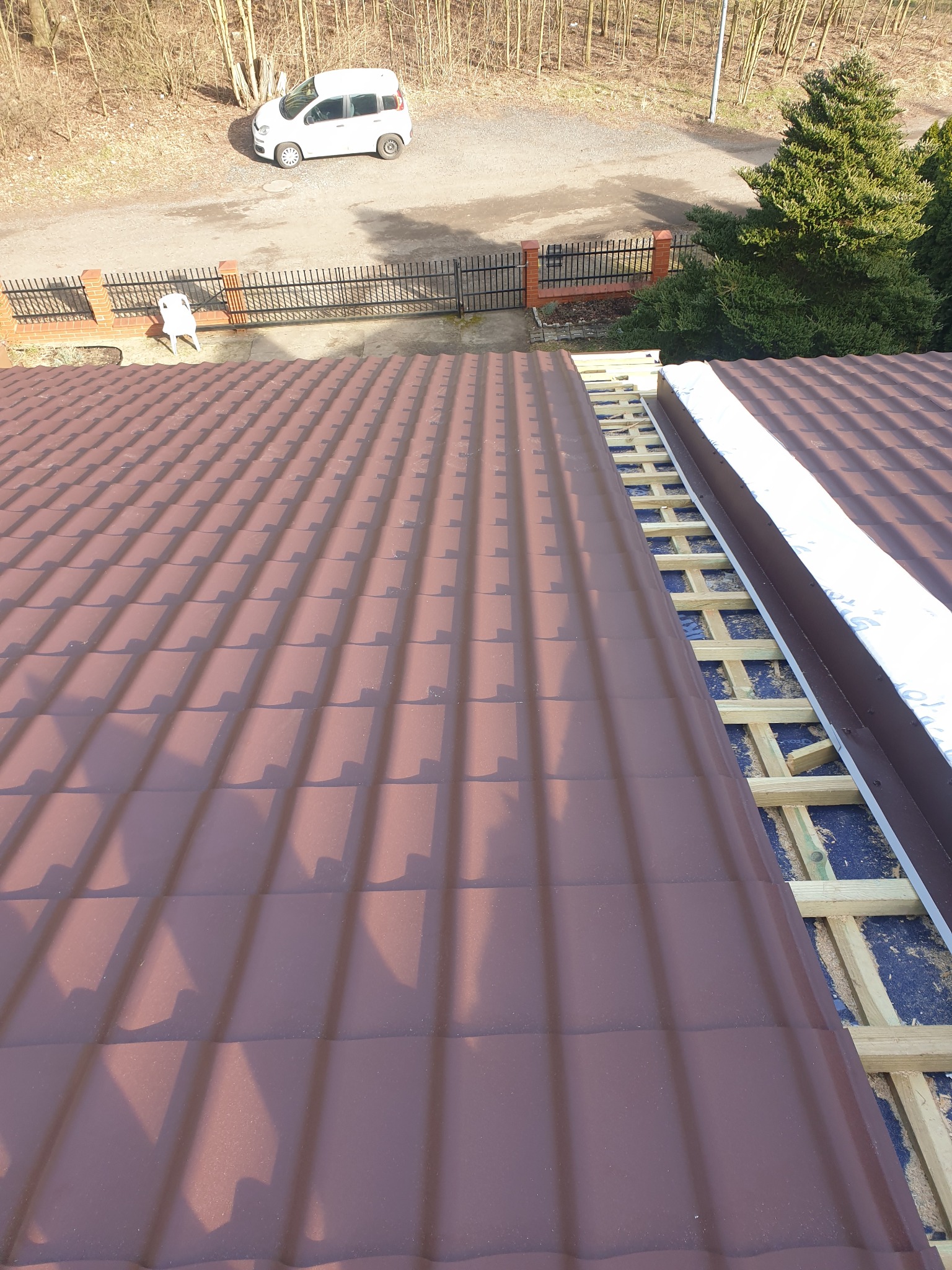 Montaż brązowej blachodachówki na drewnianej konstrukcji dachu. Część dachu jeszcze bez pokrycia. Widok z góry na budynek i otoczenie.