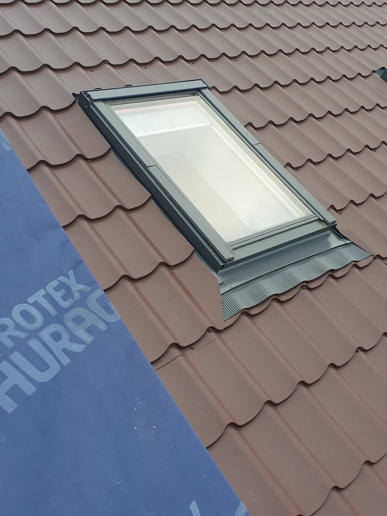Brązowy dach z blachy trapezowej z zamontowanym oknem dachowym. Fragment membrany dachowej widoczny w lewym dolnym rogu kadru.