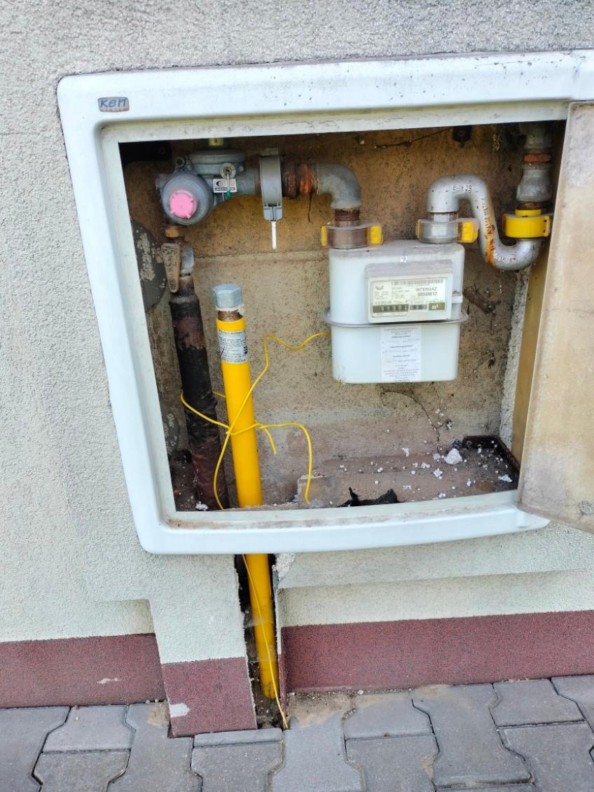 Zewnętrzna szafka gazowa z licznikiem i żółtą rurą gazową wchodzącą w ziemię, widok z bliska na instalację przy budynku.