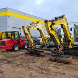 Żółto-szare mini koparki i wywrotki różnych marek stojące na placu budowy, z budynkiem w tle. Widoczne detale: AMMANN YANMAR, WACKER NEUSON, SCHAFFER.