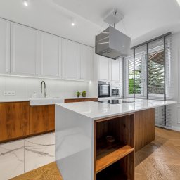 Sebastian Pietrak - ProperPlace Sp. z o.o. - Nowoczesna kuchnia z wyspą: białe szafki, drewniane akcenty i marmurowa podłoga. Okno z żaluzjami wpuszcza światło na parkiet ułożony w jodełkę.