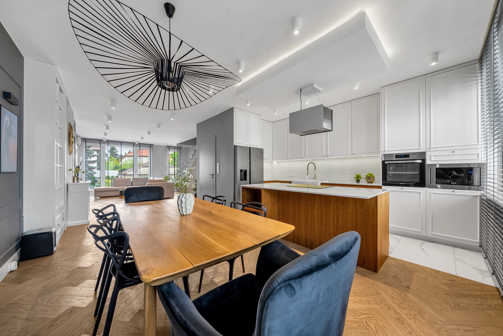 Jasne, przestronne wnętrze z drewnianym stołem i krzesłami, otwarte na kuchnię z wyspą. Charakterystyczny, designerski żyrandol nad stołem. Parkiet w jodełkę.