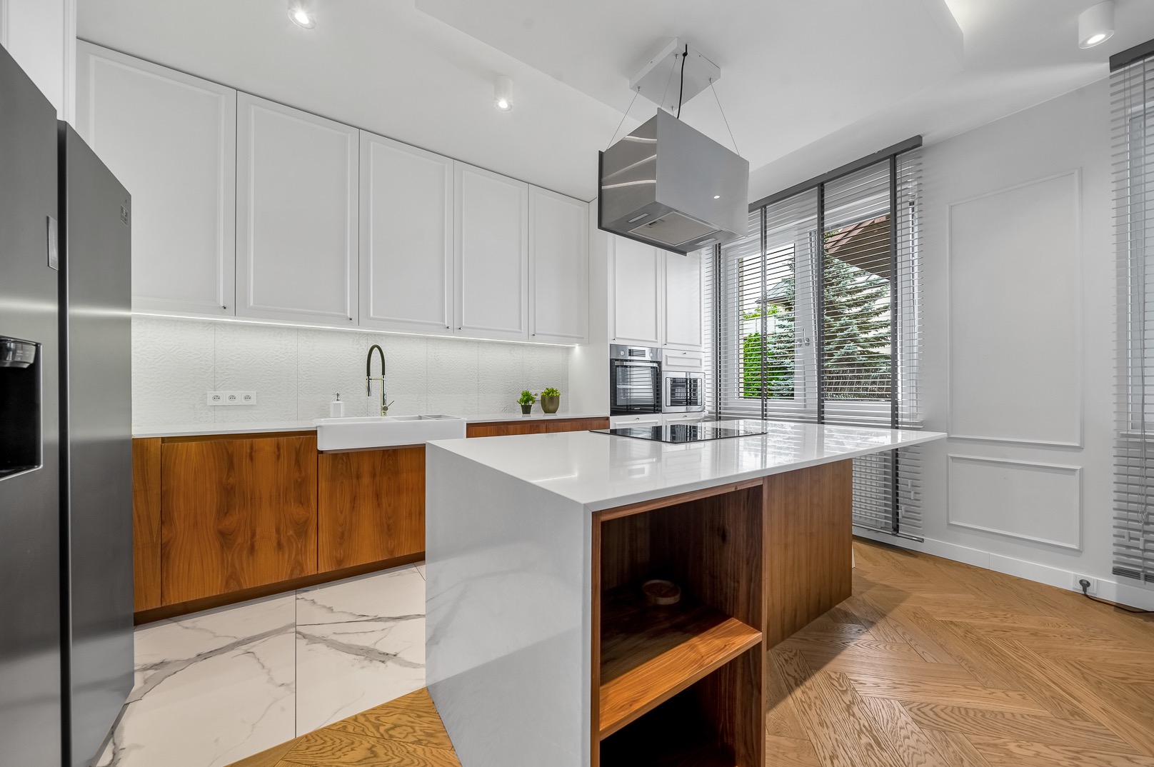 Nowoczesna kuchnia z wyspą: białe szafki, drewniane akcenty i marmurowa podłoga. Okno z żaluzjami wpuszcza światło na parkiet ułożony w jodełkę.