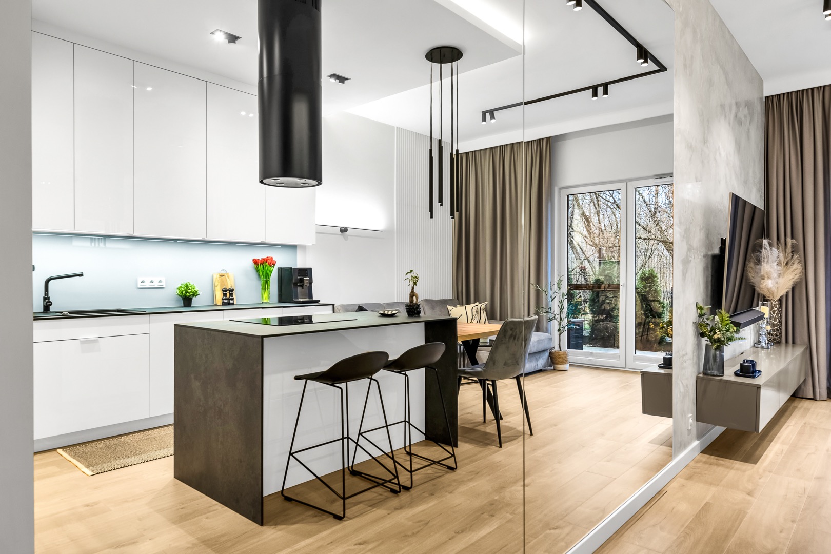 Nowoczesne wnętrze: biała kuchnia z wyspą, czarne hokery i okap, salon z tv za szklaną ścianą, drewniana podłoga. Minimalistyczny design, naturalne światło.