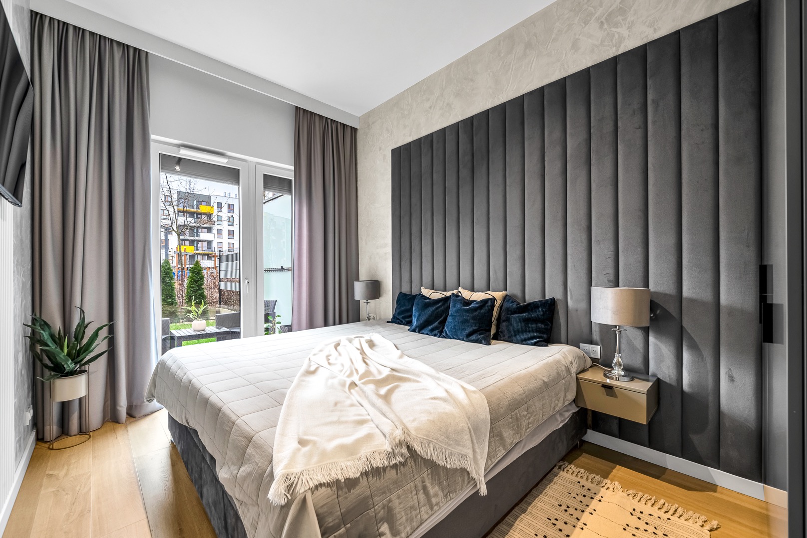 Sypialnia z dużym łóżkiem, wezgłowiem z paneli, balkonem i widokiem na osiedle. Nowoczesny design, stonowana kolorystyka, drewniana podłoga, roślina w doniczce.