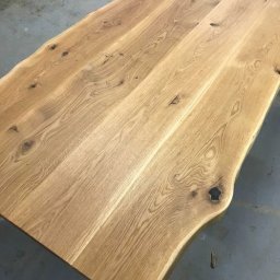 drewniane.tech - Blat z litego drewna dębowego z naturalną krawędzią, widoczne słoje i sęki, wykończony na gładko, na tle szarej podłogi. Detal stolarski.