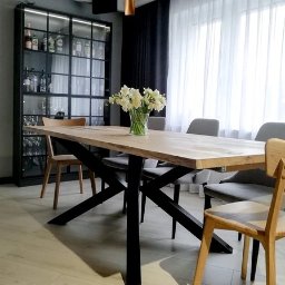 drewniane.tech - Drewniany stół jadalny z czarnymi nogami w kształcie litery X, otoczony krzesłami. Na stole wazon z białymi kwiatami, w tle przeszklona szafa z oświetleniem.