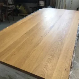 Blat drewniany z widocznym usłojeniem, zabezpieczony folią. W tle warsztat stolarski z narzędziami i akcesoriami.