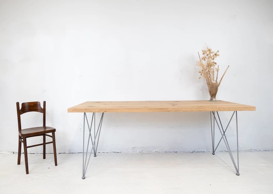 Stół z blatem z jasnego drewna o prostych krawędziach i metalowych nogach w stylu hairpin, obok krzesło z ciemnego drewna i wazon z suszonymi kwiatami.