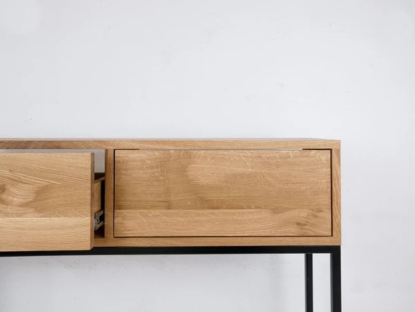 Minimalistyczna komoda z dębowego drewna na czarnych, metalowych nogach. Jedna szuflada wysunięta, ukazująca prowadnice. Jasne, naturalne wykończenie drewna.