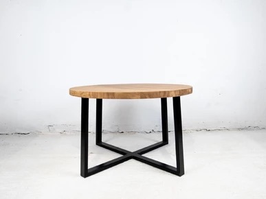 Okrągły stolik kawowy z jasnego drewna na czarnych, metalowych nogach w kształcie litery X. Minimalistyczny design na tle białej ściany.