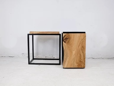 Dwa stoliki kawowe: jeden z blatem i czarną, metalową ramą, drugi pełny, z wyraźnym usłojeniem drewna i czarnym obramowaniem, na tle białej ściany.