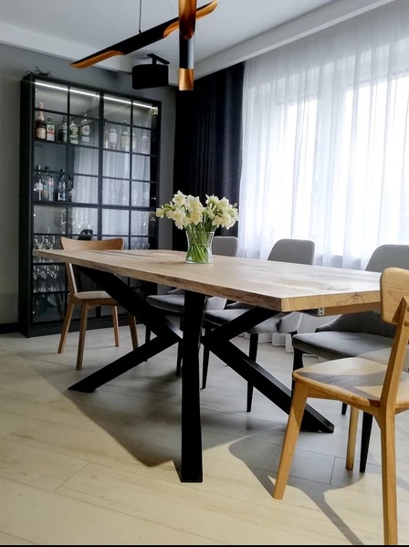 Drewniany stół jadalny z czarnymi nogami w kształcie litery X, otoczony krzesłami. Na stole wazon z białymi kwiatami, w tle przeszklona szafa z oświetleniem.