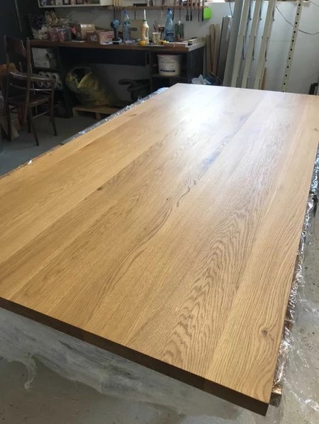 Blat drewniany z widocznym usłojeniem, zabezpieczony folią. W tle warsztat stolarski z narzędziami i akcesoriami.