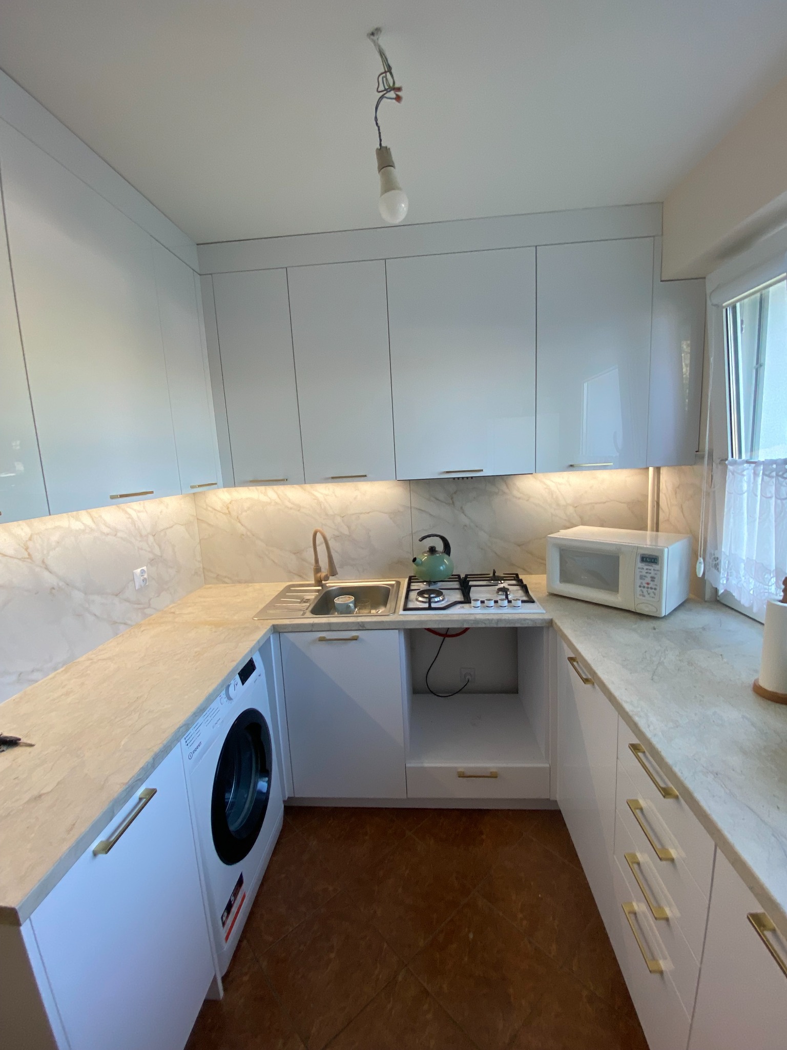 Biała kuchnia z marmurowym blatem, złotymi uchwytami i pralką w zabudowie. Nad blatem włączone oświetlenie LED. Widoczny fragment okna z firanką.