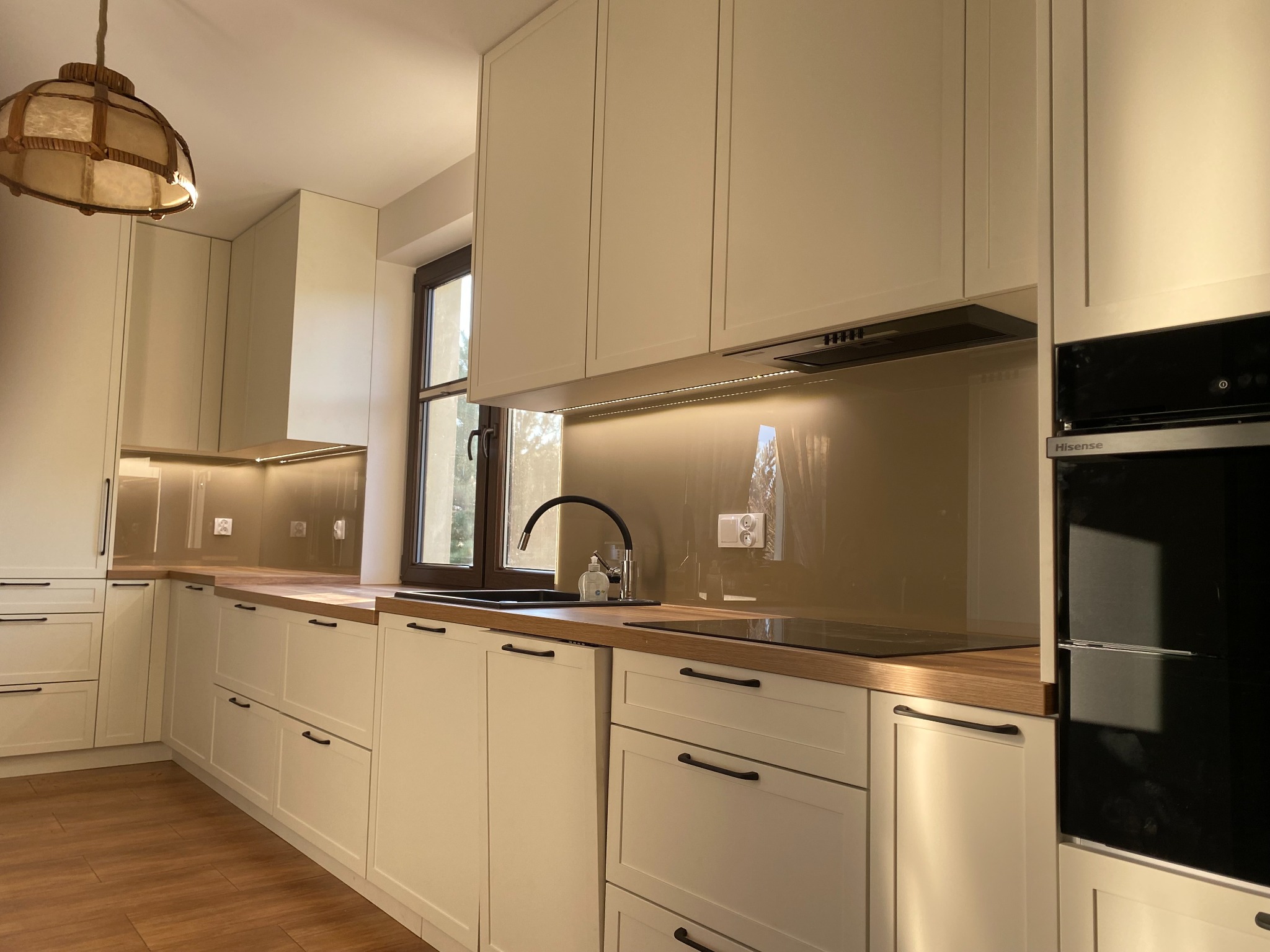 Jasna kuchnia na wymiar z drewnianym blatem i czarnymi uchwytami. Widoczne okno z brązową ramą i lampa wisząca z abażurem w stylu retro. Całość w ciepłej tonacji.