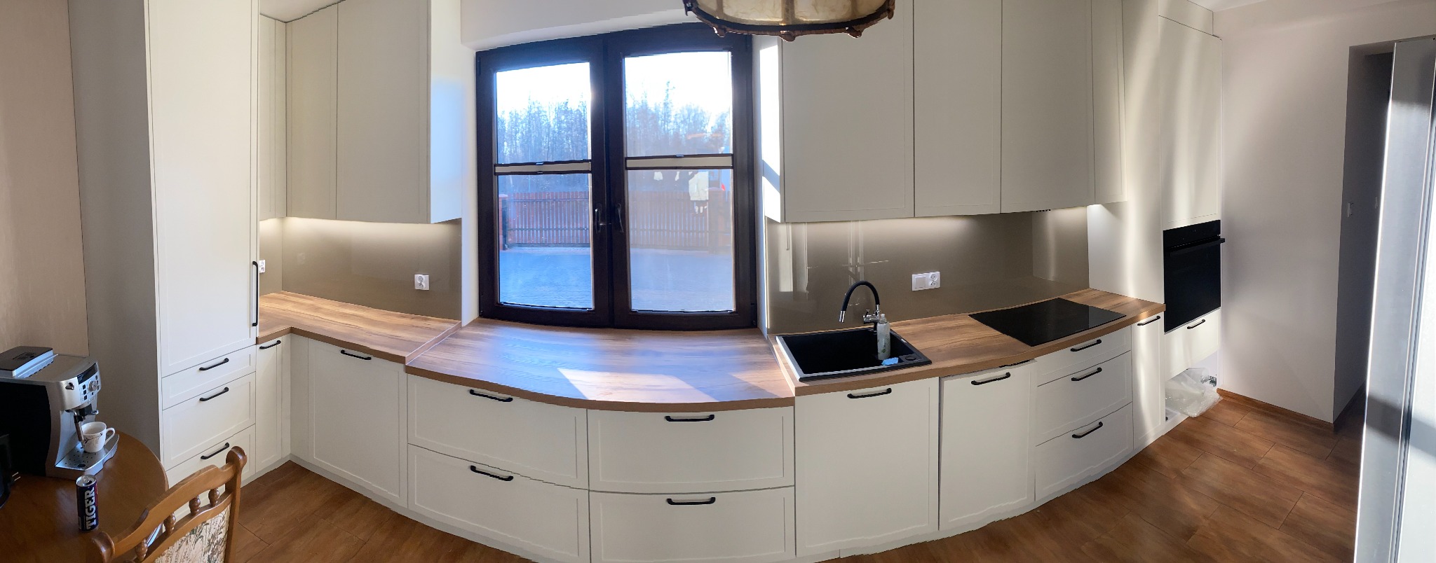 Jasna kuchnia na wymiar z drewnianym blatem, czarnym zlewem i oknem z widokiem na zewnątrz. Uwagę przykuwa drewniany blat pod oknem.