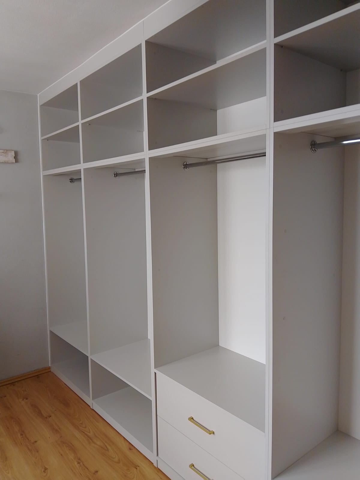 Biała szafa wnękowa z półkami i drążkami na ubrania, z widocznymi szufladami z złotymi uchwytami. Jasne wnętrze, drewnopodobna podłoga.