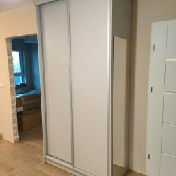 ZAKŁAD STOLARSKI ANDRZEJ CIELUCH - Jasna szafa wnękowa z przesuwnymi drzwiami i lustrem bocznym, na tle wykończonego wnętrza z drewnopodobną podłogą i widokiem na pokój z oknem.