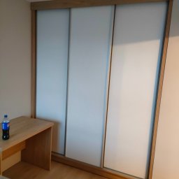 ZAKŁAD STOLARSKI ANDRZEJ CIELUCH - Jasna szafa wnękowa z przesuwnymi drzwiami i drewnianą ramą, z blatem po lewej stronie, na którym stoi butelka napoju. Minimalistyczny design.