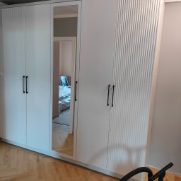 ZAKŁAD STOLARSKI ANDRZEJ CIELUCH - Biała szafa wnękowa z lustrem i frezowanymi frontami, czarne podłużne uchwyty, parkiet w jodełkę, widoczny odkurzacz. Wnętrze pokoju widoczne w lustrze.