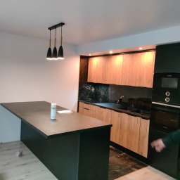 ZAKŁAD STOLARSKI ANDRZEJ CIELUCH - Nowoczesna kuchnia z wyspą: drewniane fronty szafek, czarny blat i marmurowy backsplash. Nad wyspą wiszą trzy czarne lampy.