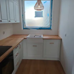 Jasna kuchnia z drewnianym blatem i białymi szafkami, okno zabezpieczone folią, lampa wisząca. Widok na fragment płyty grzewczej i piekarnika.