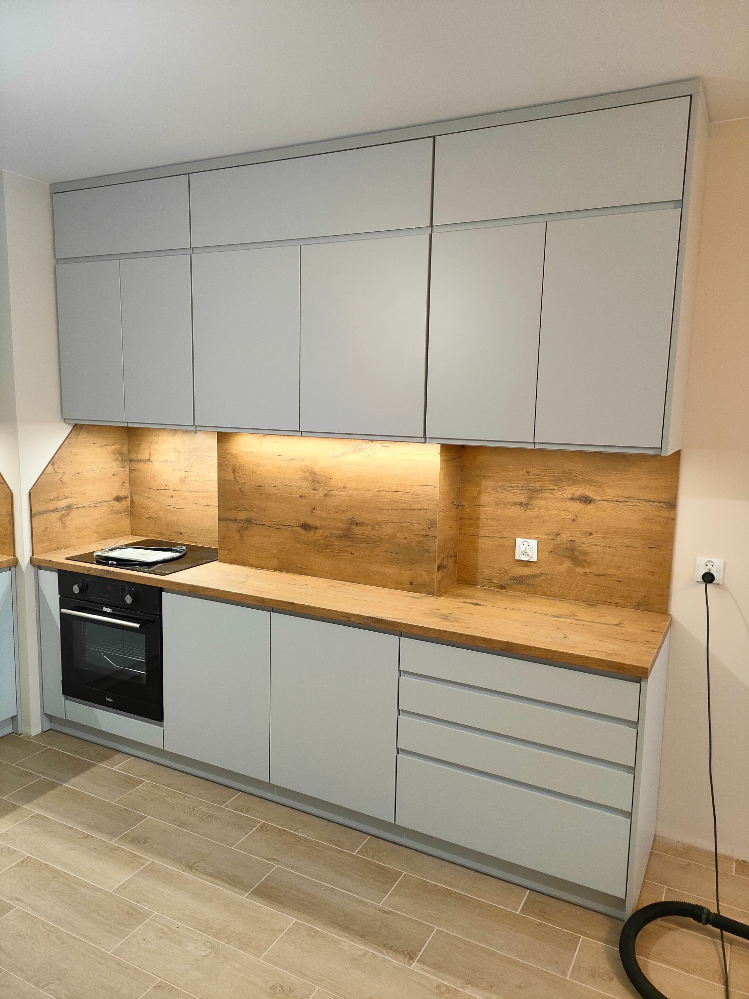 Jasnoszara zabudowa kuchenna z drewnianym blatem i panelem. Oświetlenie podszafkowe podkreśla naturalny dekor drewna. Widoczna płyta indukcyjna i piekarnik.