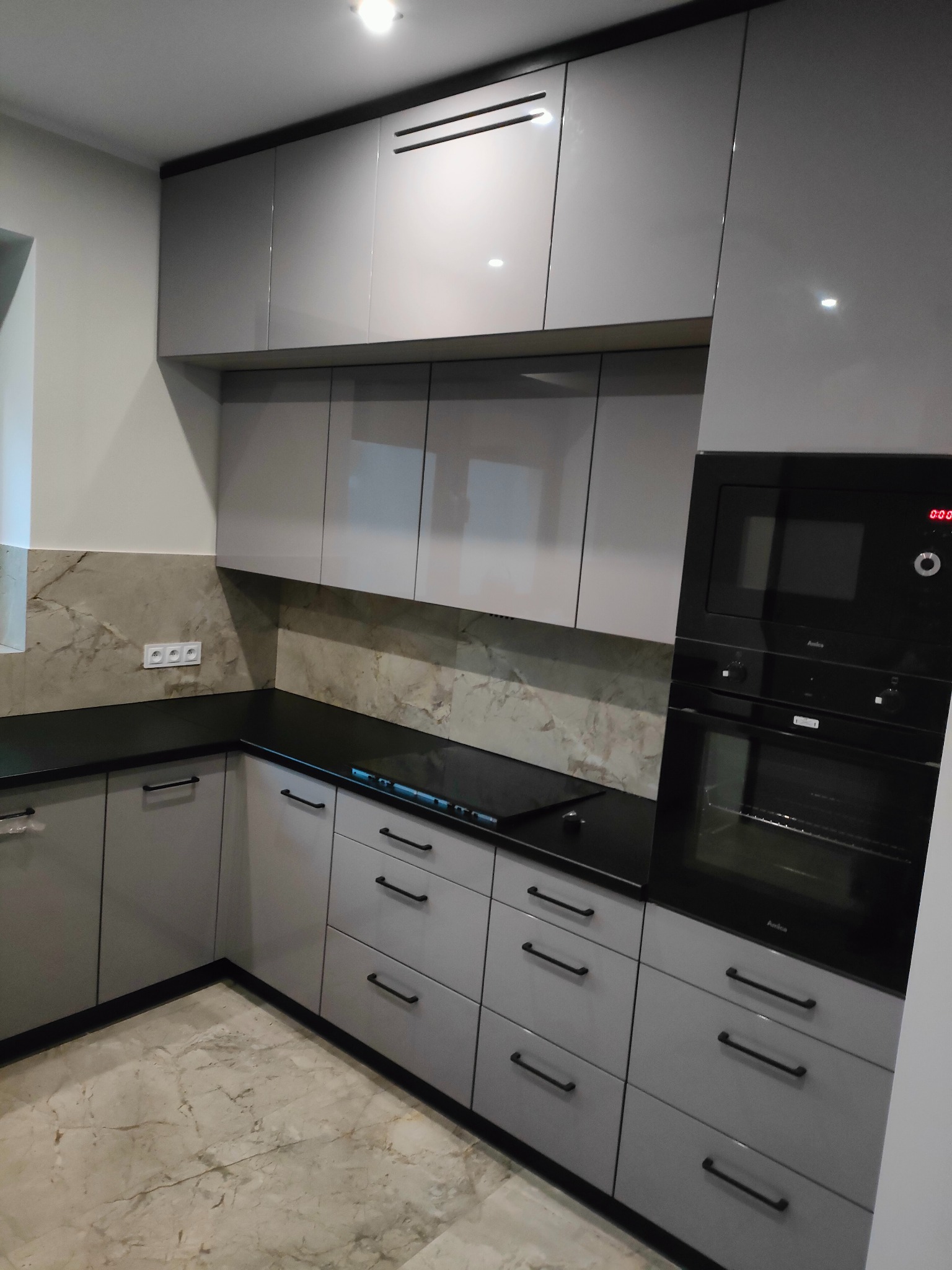 Szare, lakierowane meble kuchenne z czarnym blatem i sprzętem AGD w Twardogórze. Górne szafki z poziomymi uchwytami, marmurowa ściana nad blatem.