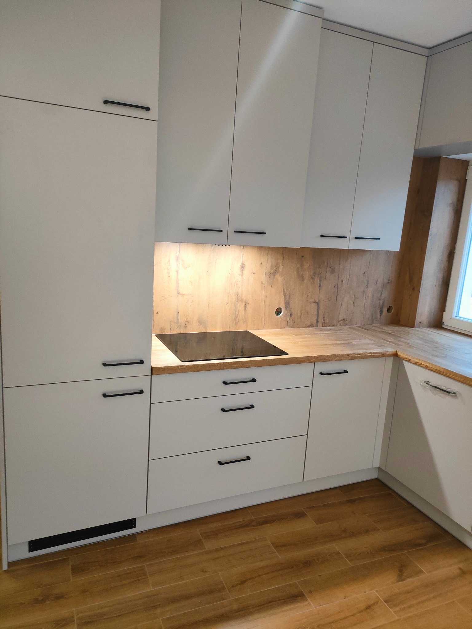 Biała zabudowa kuchenna z drewnianym blatem i czarnymi uchwytami. Widoczna płyta indukcyjna i drewniana ściana nad blatem. Ujęcie z perspektywy podłogi.