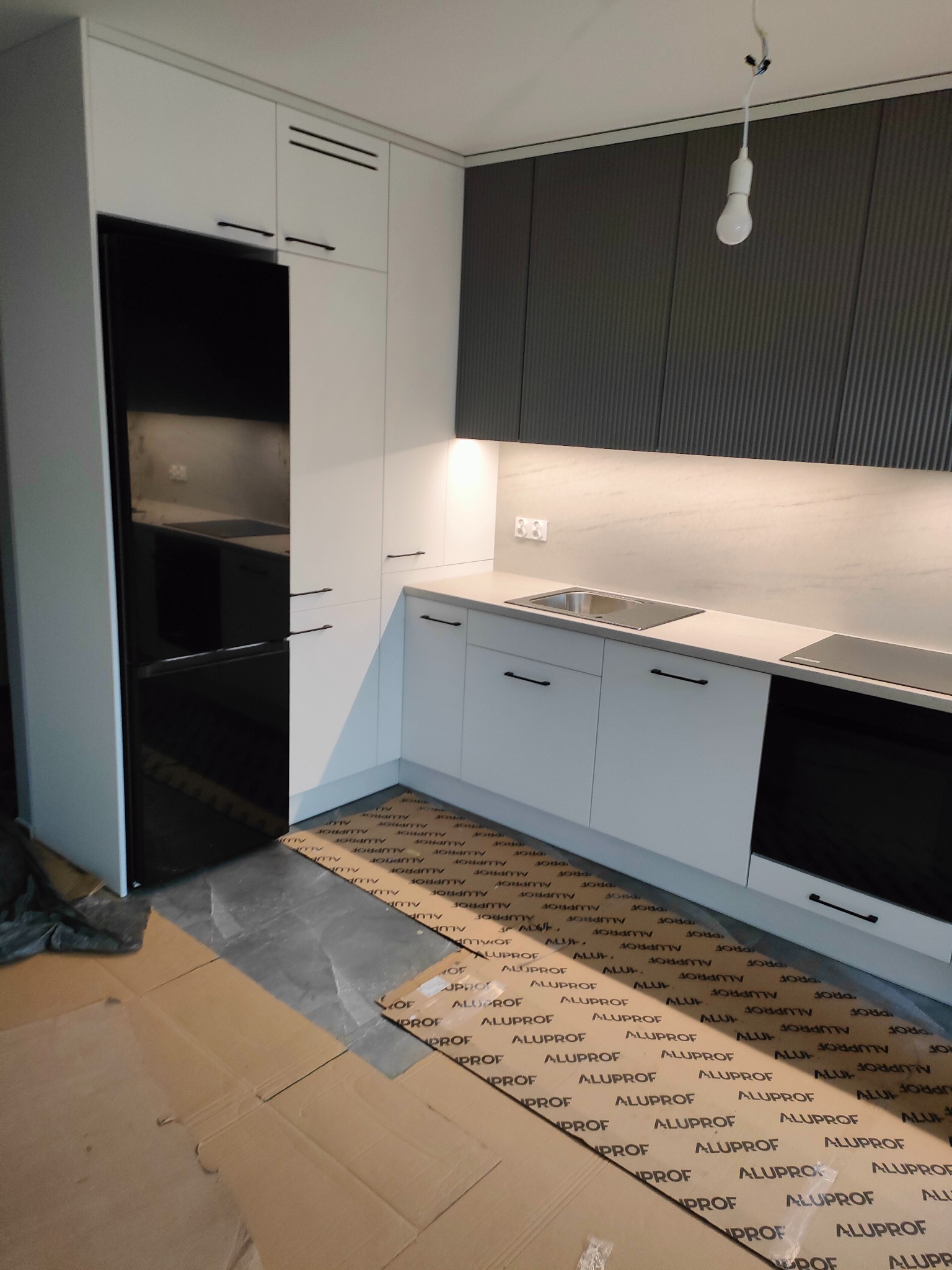 Nowoczesna kuchnia z białymi i grafitowymi szafkami, czarnym sprzętem AGD i podłogą zabezpieczoną kartonem. Minimalistyczne uchwyty, oświetlenie punktowe.