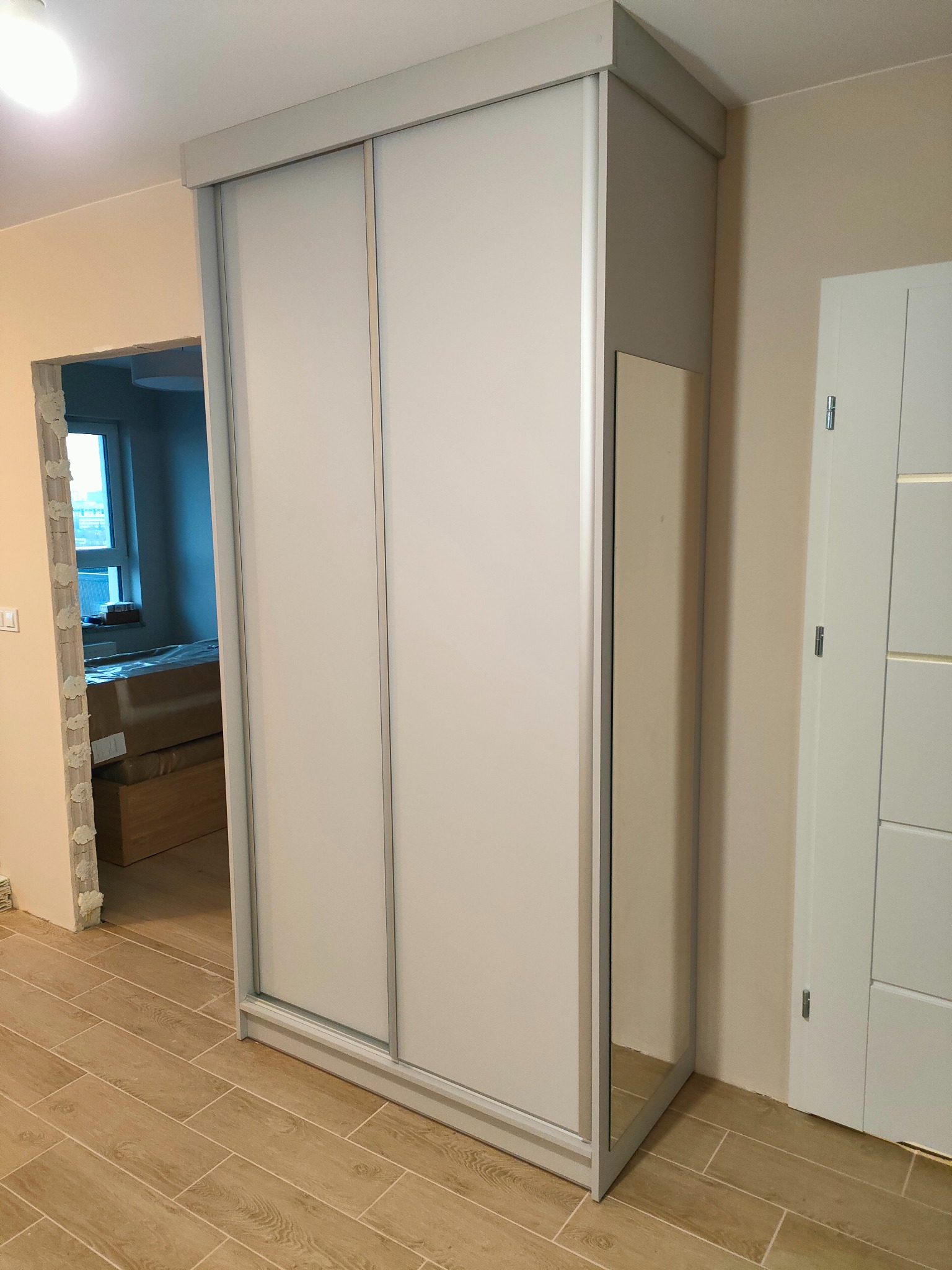 Jasna szafa wnękowa z przesuwnymi drzwiami i lustrem bocznym, na tle wykończonego wnętrza z drewnopodobną podłogą i widokiem na pokój z oknem.